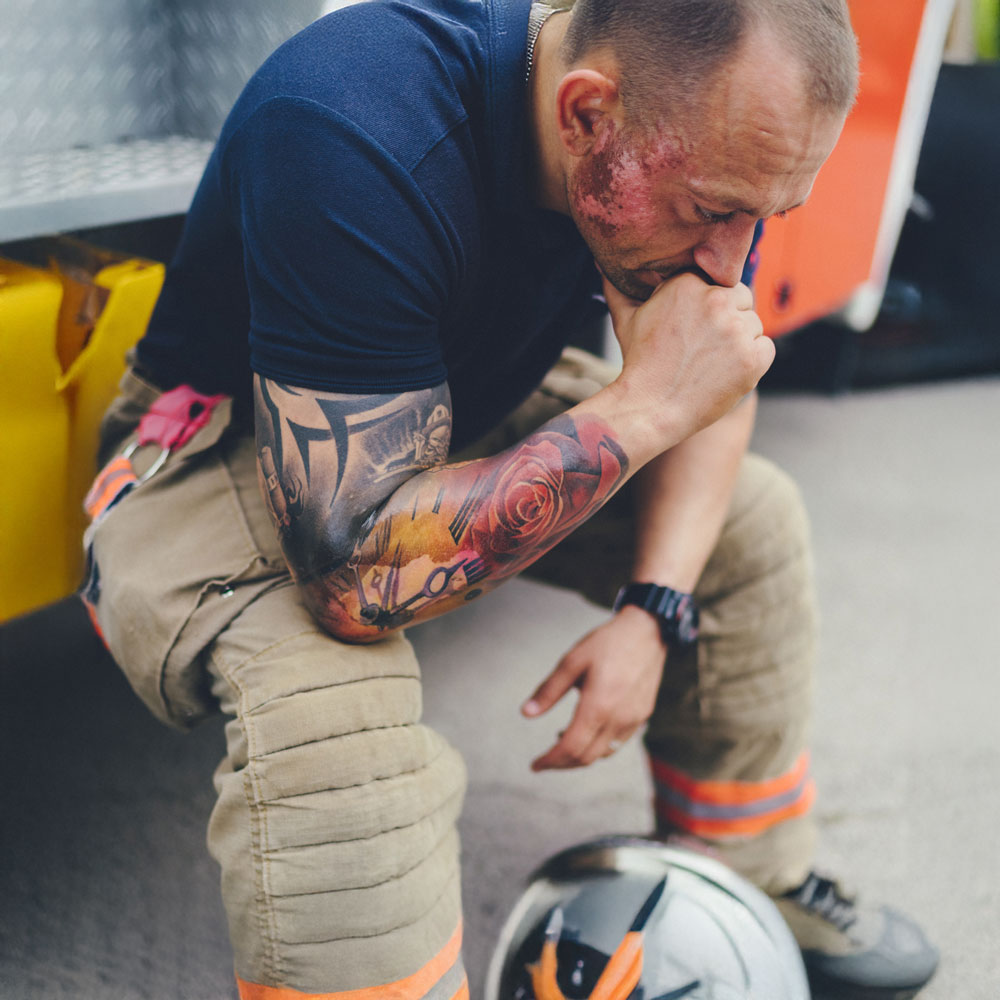 Depressed firefighter with facial burns sitting on the side of a firetruck after a failed attempt to rescue a child from a burning building.