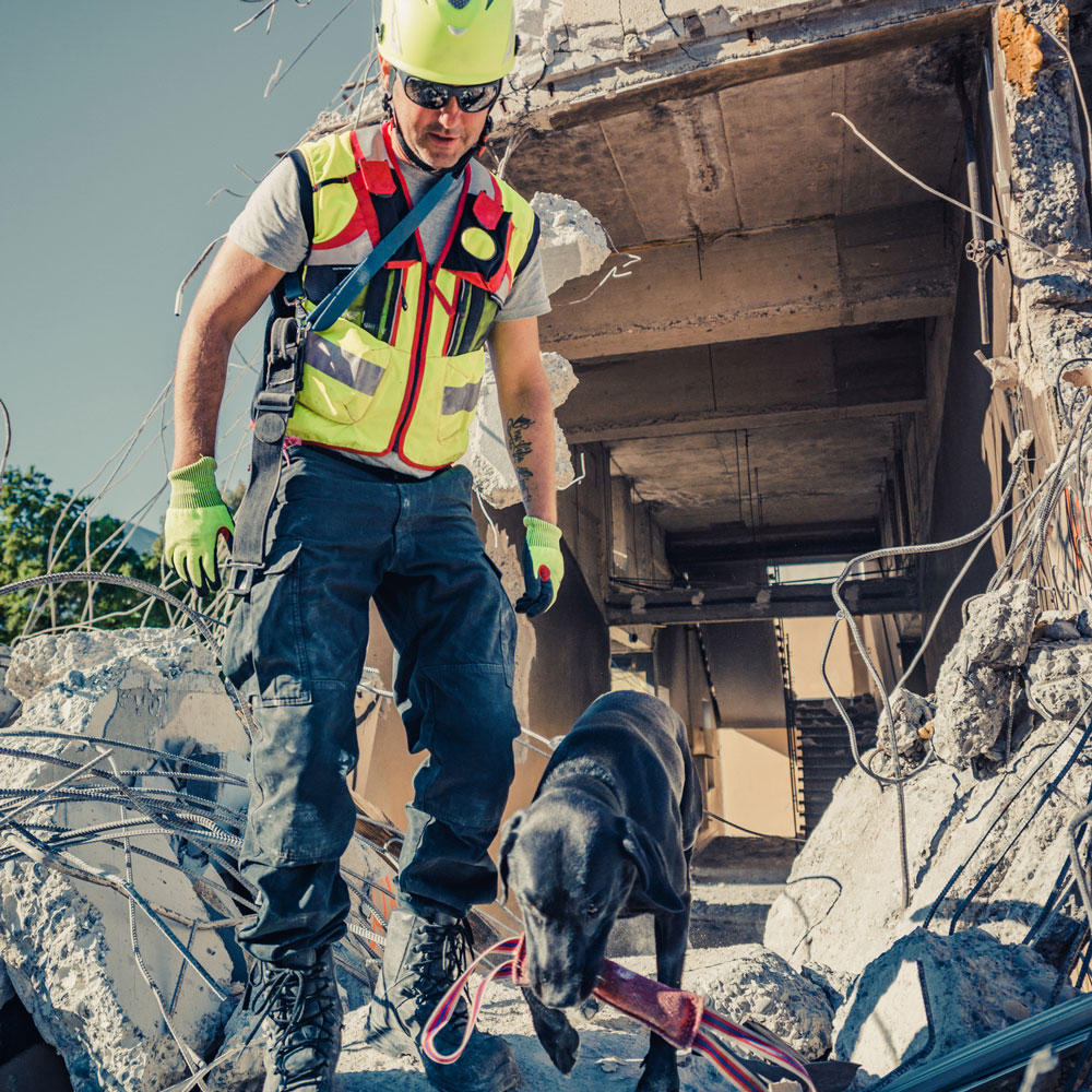 Search and rescue worker with a cadaver dog walking on the remains of a crumbled building, highlighting the need for first responder PTSD and mental health treatment programs at Deer Hollow.