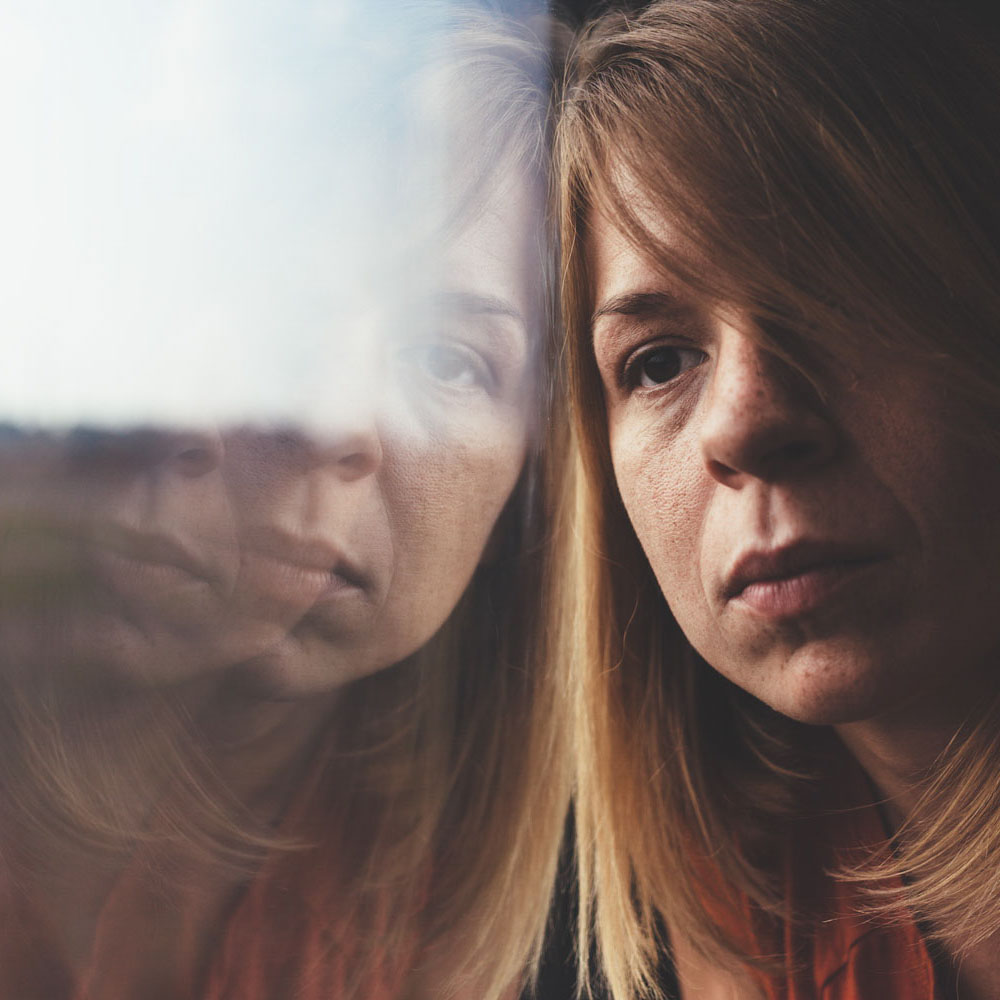 Depressed nurse suffering from addiction and unresolved trauma, staring out a window, highlighting the need for comprehensive treatment at addiction rehabilitation centers like Deer Hollow to address both PTSD and addiction for lasting recovery.