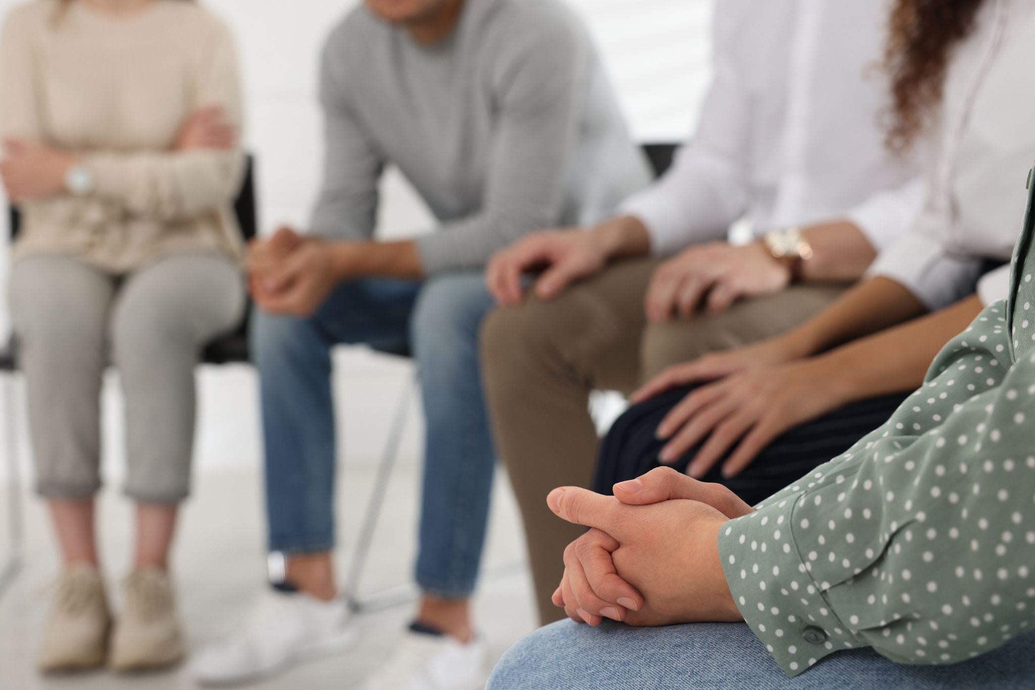 A close-up of a person's clasped hands with other people sitting in a therapy group out of focus in the background.