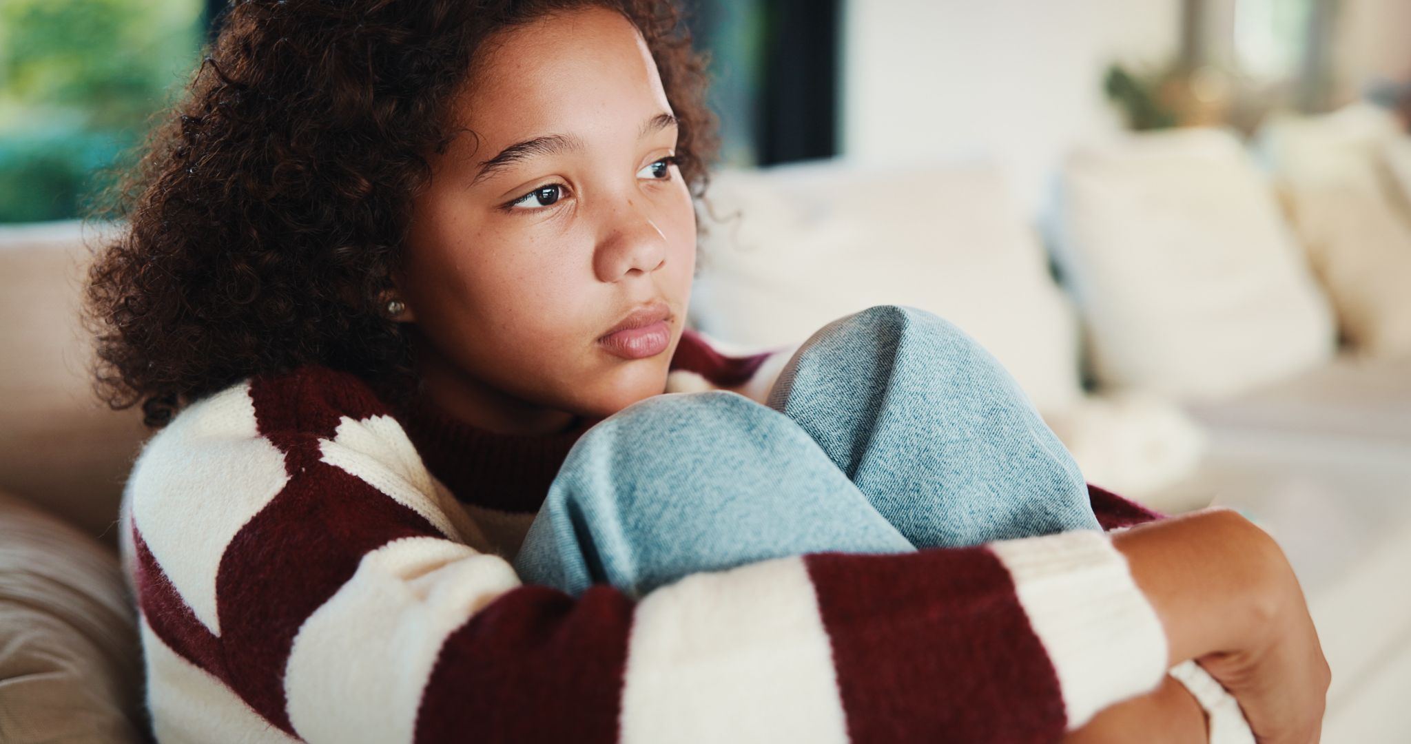 Young girl sitting on a couch hugging her knees with a thoughtful, pensive expression.