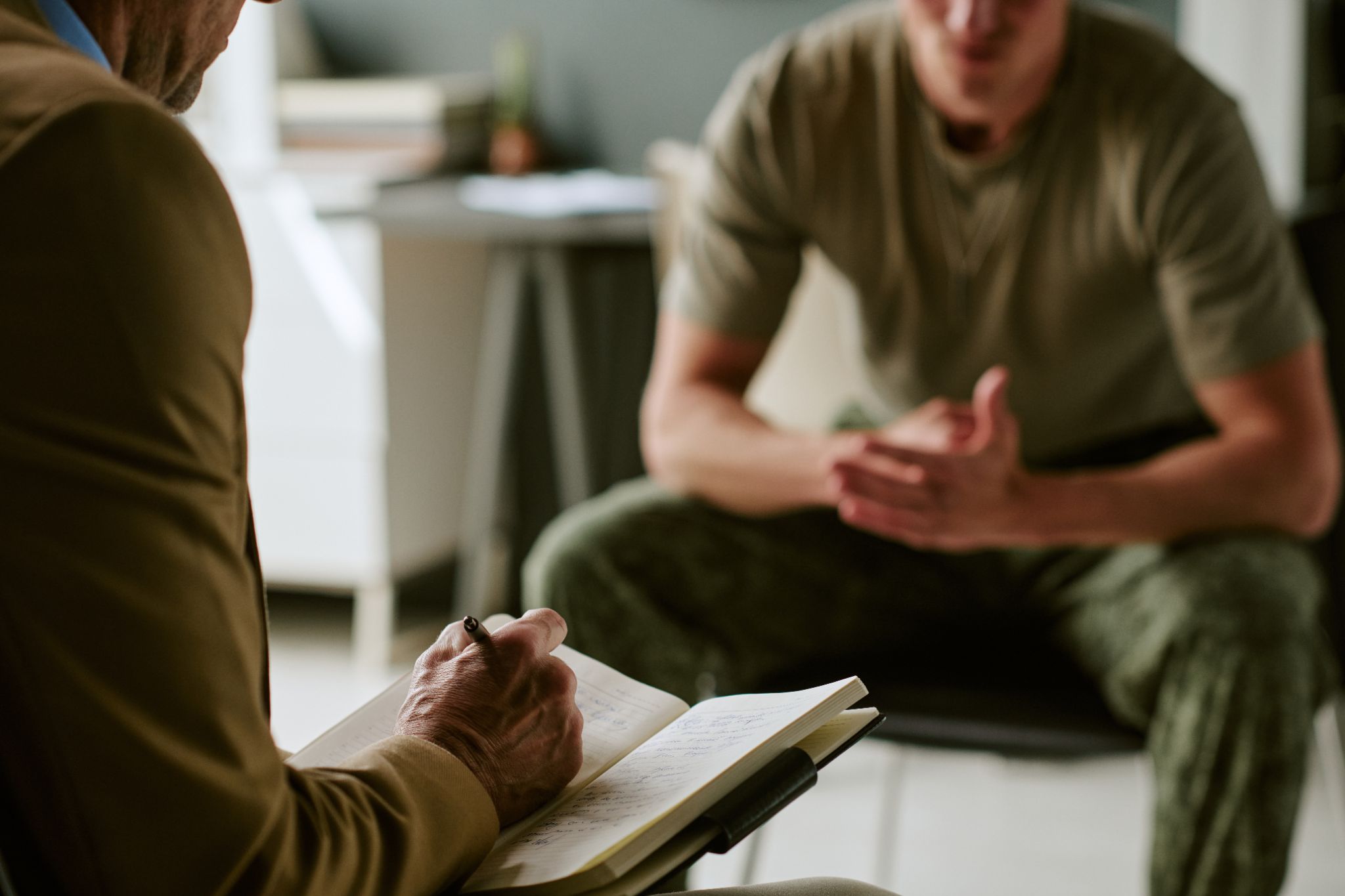 Therapist taking notes in a notebook while speaking with a military service member in camouflage pants during a session.
